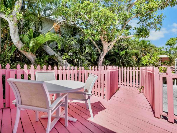 A tropical garden patio with pink picket fence, a white lounge chair and table, and lush palms under blue sky.