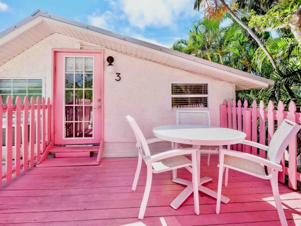 A pink house deck with a round table, chairs, picket railing, and lush palm trees under a blue sky.