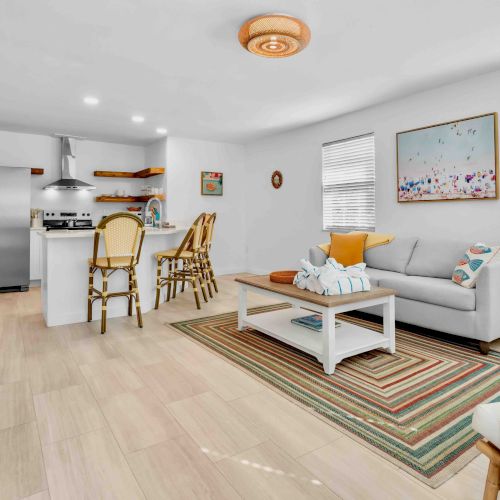 Bright living room with sofa, coffee table, rug, and a kitchen area with fridge, stove, and bar stools.