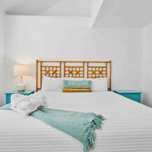 A bright bedroom with a white bed, turquoise side tables, a bamboo headboard, a lamp, and shutters on the wall.
