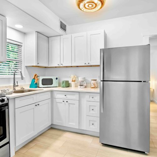 Bright modern white kitchen with cabinets, sink, stove, microwave, coffee maker, and refrigerator, with a ceiling light overhead.