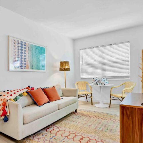 Bright living room with beige sofa, colorful pillows, framed art, lamp, two chairs, TV, and white window blinds.