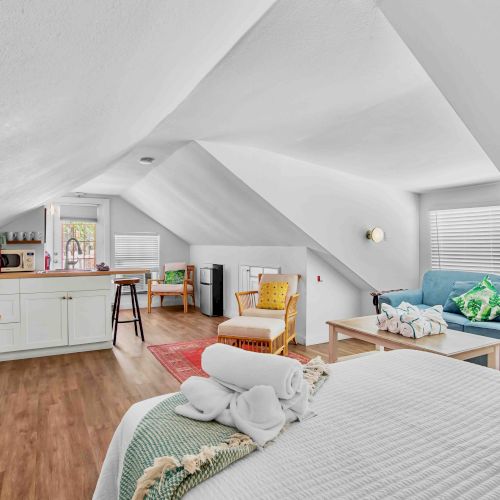 A bright attic living room with a blue sofa, kitchenette cabinets, TV, coffee table, chairs, and white sloped ceilings.