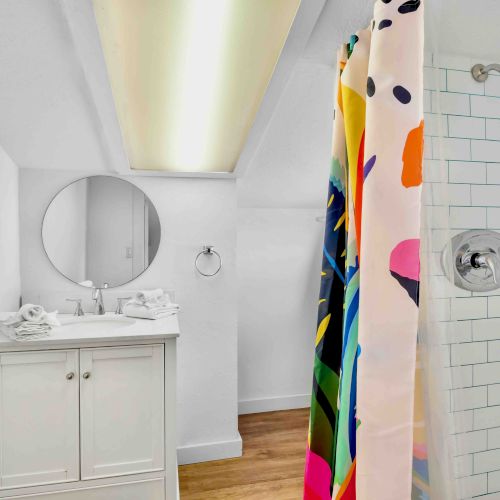 Bright bathroom with white vanity and round mirror, plus a tiled shower with colorful curtain and chrome showerhead.