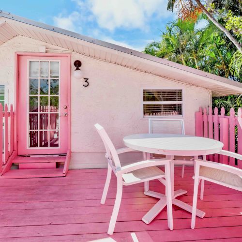 A pink porch outside a small house with a round table, chairs, and tall palm trees in bright daylight.