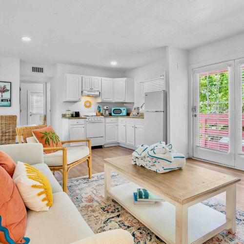 Bright open-plan living room and kitchen with white cabinets, windows with blinds, and cozy couches and decor inside.
