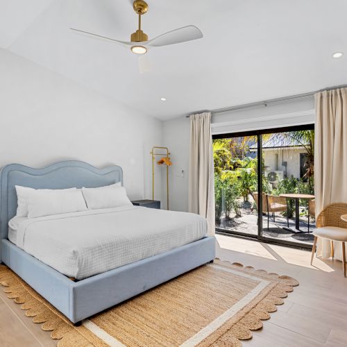 A modern bedroom with a blue bed, ceiling fan, beige curtains, and glass doors opening to a lush garden patio.