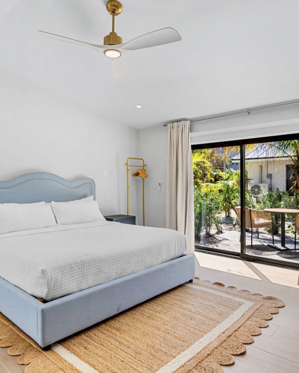 A modern bedroom with a blue bed, ceiling fan, beige curtains, and glass doors opening to a lush garden patio.
