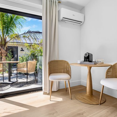 Sunny patio dining nook with rattan chairs, a coffee machine, wall AC, and sliding doors opening to palms outside.