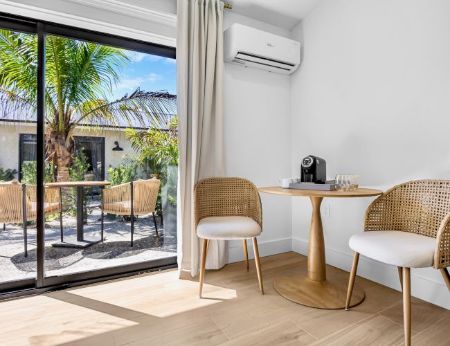 Sunny patio dining nook with rattan chairs, a coffee machine, wall AC, and sliding doors opening to palms outside.
