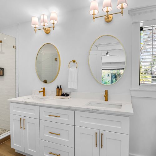 Bright modern bathroom with a white double vanity, gold fixtures, oval mirrors, pendant vanity lights, and a window with blinds.
