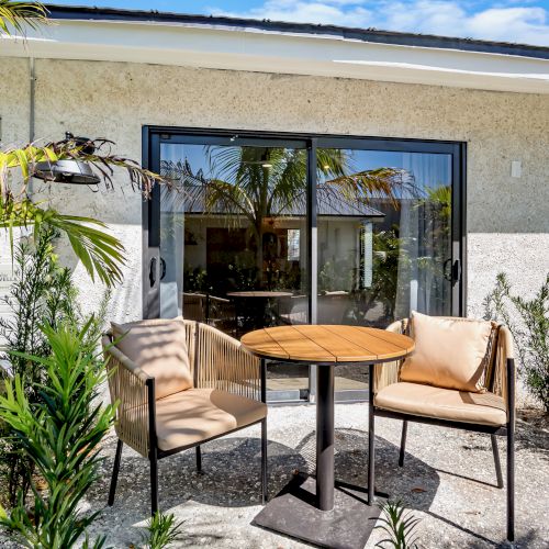 A tropical patio with cushioned chairs and a wooden table beside a wall, glass doors, lush plants, and an outdoor AC unit.