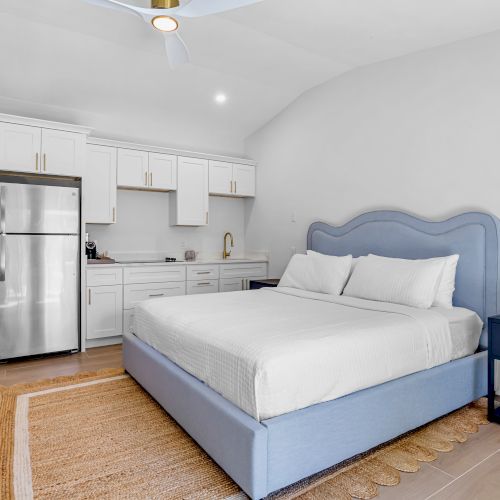Bright modern bedroom with a blue bed, white cabinetry, stainless fridge, and gold accents, plus ceiling lights.