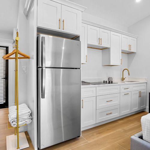 Bright kitchen with stainless double-door refrigerator, white cabinets, gold hardware, sink, and a hanging rack with clothes.