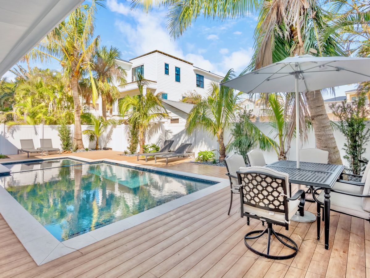 A backyard features a pool, wooden deck, patio furniture with an umbrella, and palm trees, creating a relaxing outdoor atmosphere.