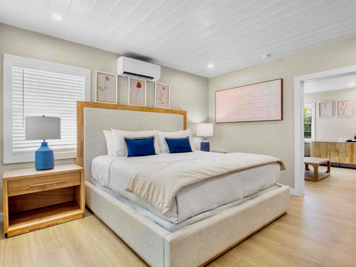 A bright modern bedroom with a large bed, bedside tables, wall art, a wall AC unit, and lamps.
