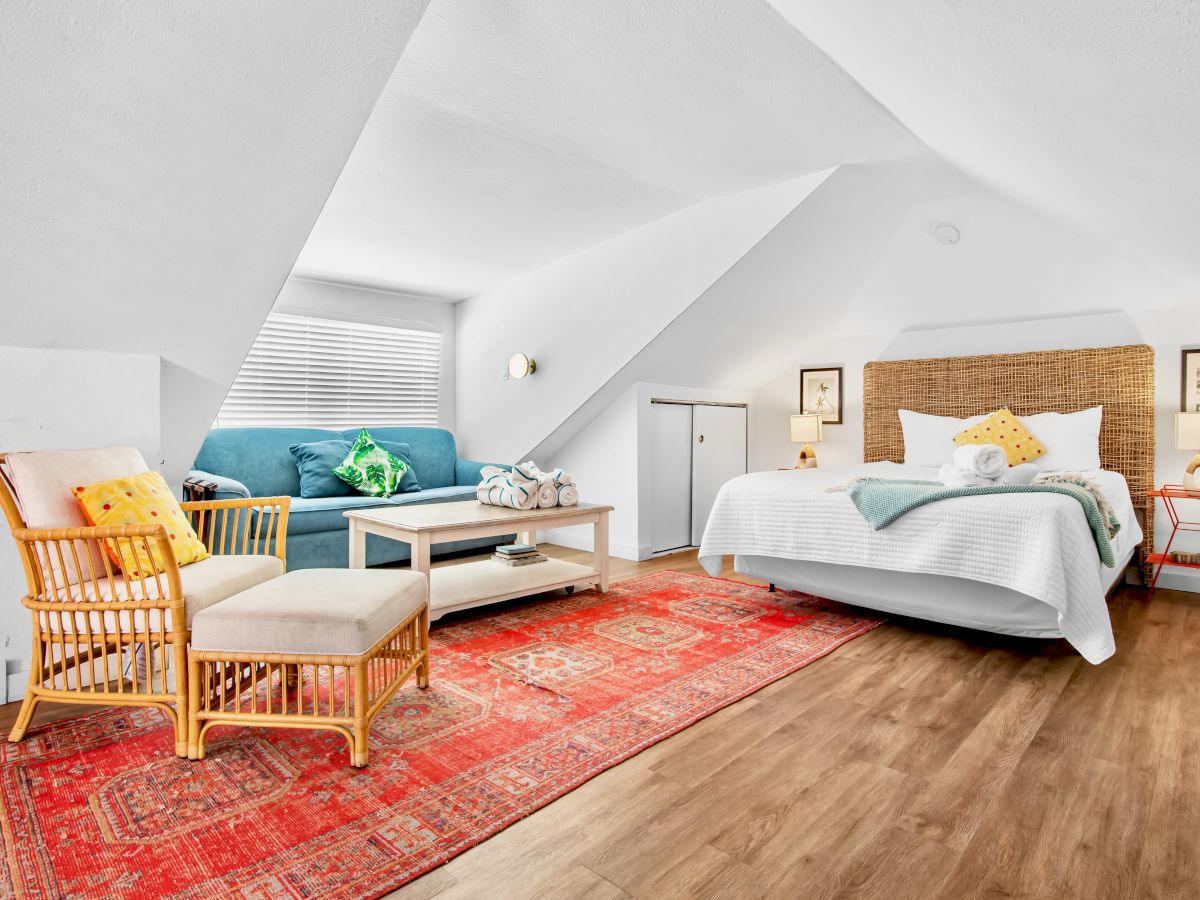 Bright loft bedroom with a sofa and bed, woven headboard, colorful rug, decorative pillows, and framed art under slanted white ceilings.