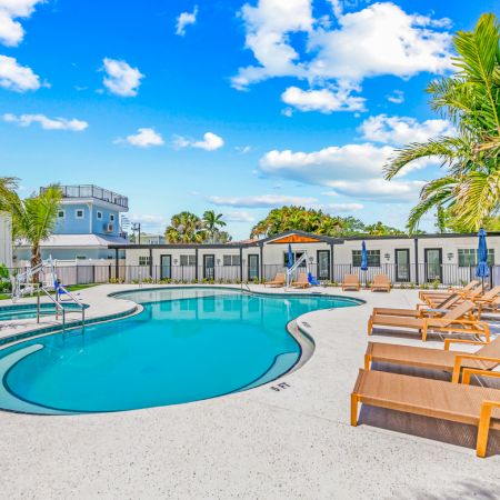 The image shows a sunny pool area with lounge chairs, umbrellas, and palm trees, set against a backdrop of a clear blue sky and clouds.