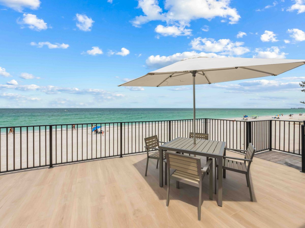 A seaside deck with a table, four chairs, and a large umbrella overlooking a calm blue ocean under a sunny sky.