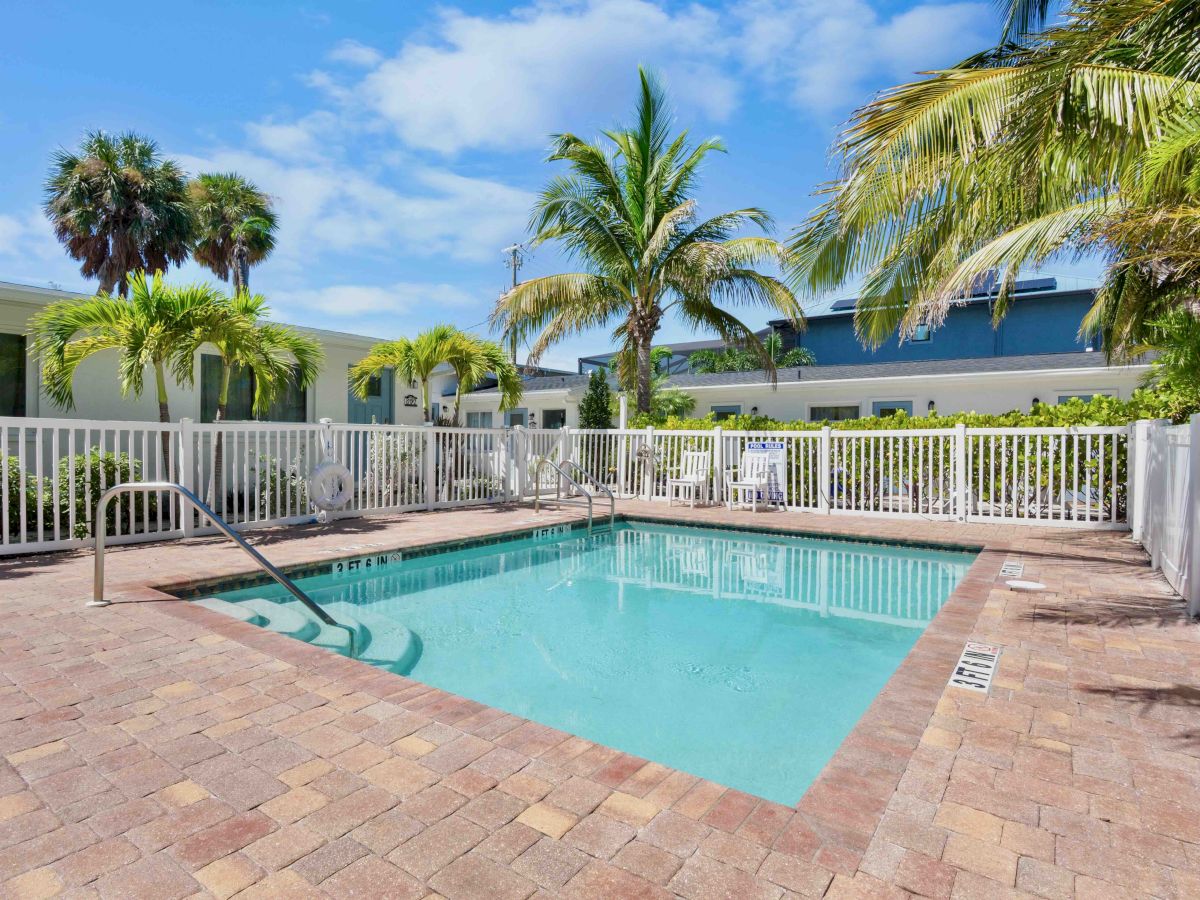 Sunny outdoor swimming pool with palm trees and a white fence, surrounded by resort buildings under a blue sky.