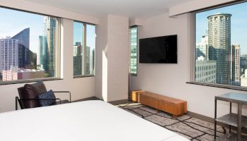 A modern hotel room with a bed, TV, and large windows showing a cityscape view of tall buildings under a clear sky.