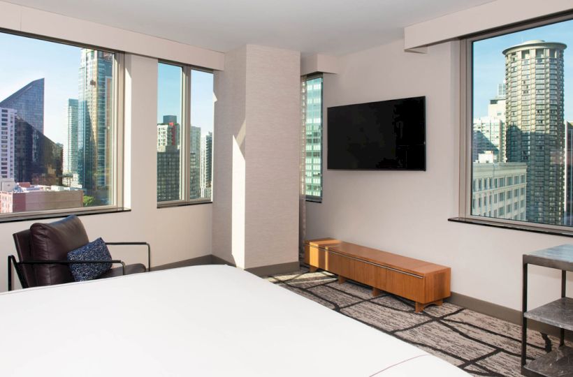 A modern hotel room with a bed, TV, and large windows showing a cityscape view of tall buildings under a clear sky.