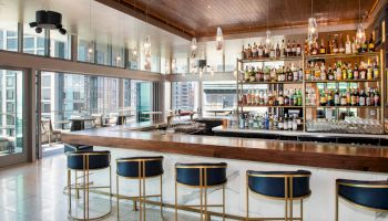 A stylish bar with high stools, a glossy wooden counter, bottles lined up, pendant lights, and large windows overlooking the city.