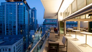 A modern urban balcony overlooks a city street with tall buildings at dusk, featuring sleek furniture and illuminated interior spaces.
