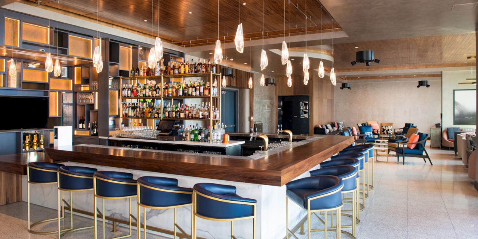A modern bar with a U-shaped counter, stylish chairs, pendant lights, and a wall stocked with various bottles in a cozy setting.