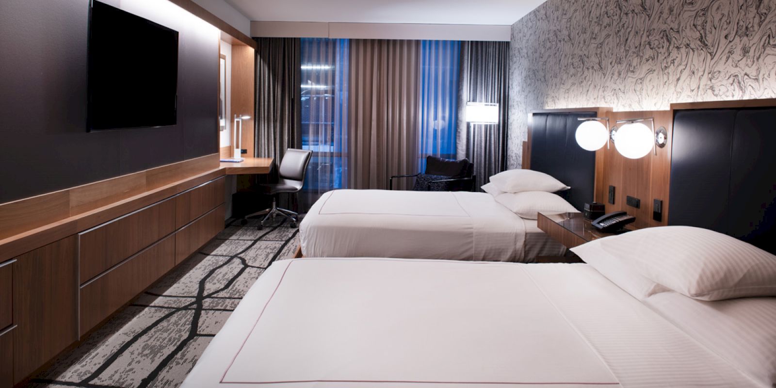 A modern hotel room with two beds, wall-mounted TV, desk, chair, and bedside lamps, featuring patterned wallpaper and curtains.