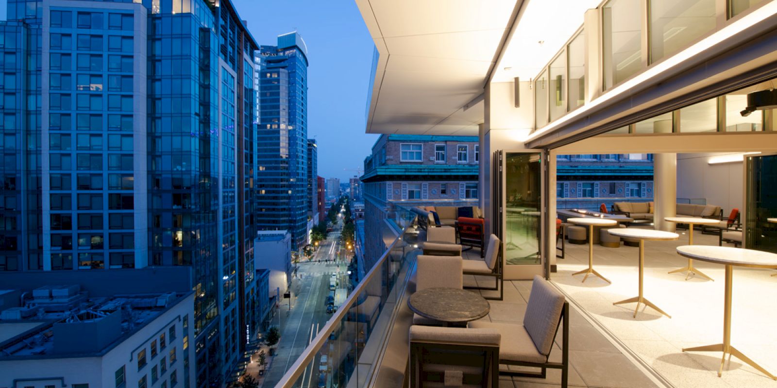 A modern rooftop patio overlooks a cityscape with tall buildings at dusk, featuring tables and chairs beside glass railings.