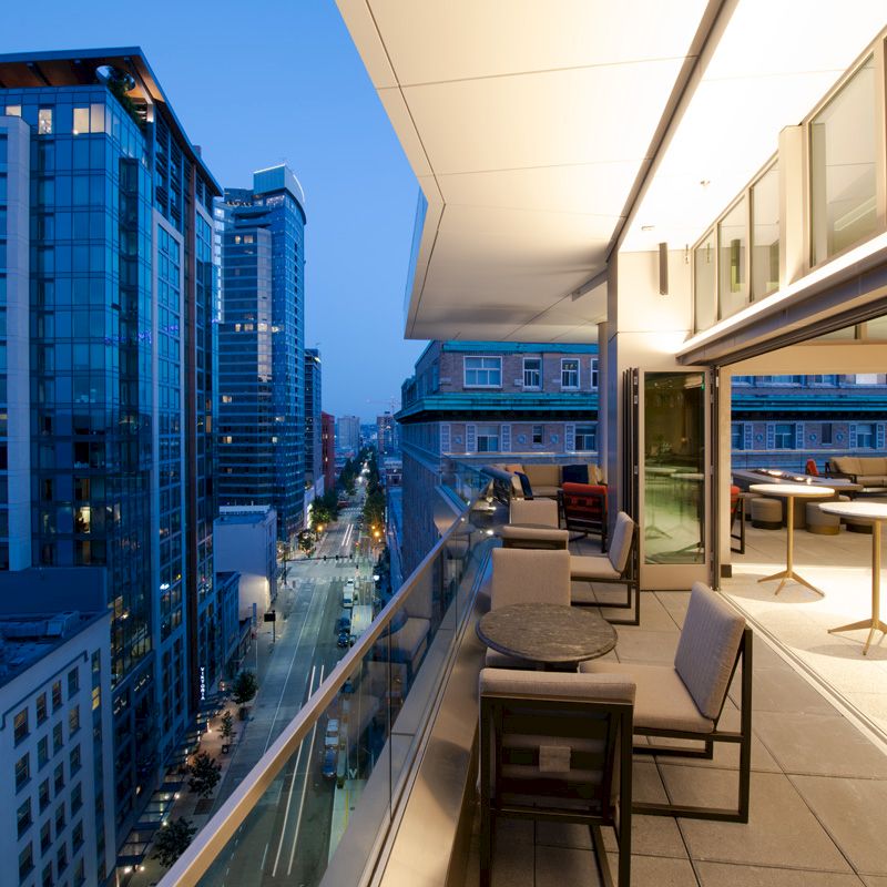 A modern urban balcony during evening with outdoor seating, overlooking city streets and high-rise buildings under a dim blue sky.