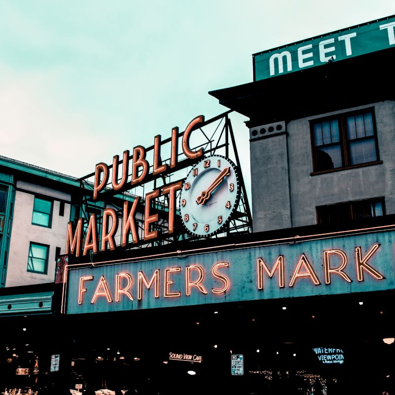 The image shows a bustling market area with neon signs reading "Public Market" and "Farmers Market" on a classic building facade.