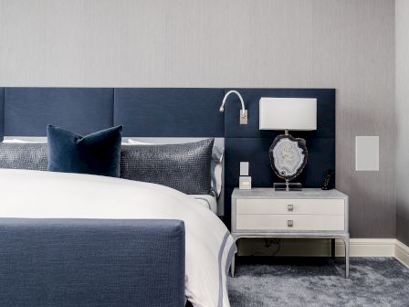 A modern bedroom with a navy bed, white bedding, gray pillows, a bedside table, lamp, and a sleek wall-mounted reading light.