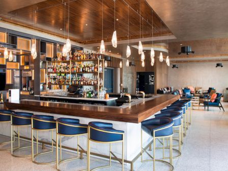 A modern bar with a wooden counter, blue chairs, and pendant lights, featuring shelves stocked with various bottles and a lounge area.