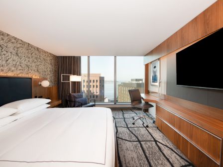 A modern hotel room features a large bed, desk, chair, wall-mounted TV, and window overlooking a distant view.
