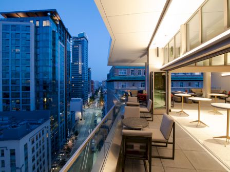 A cityscape showing a modern urban balcony with seating, adjacent to tall buildings, and illuminated by evening lights.