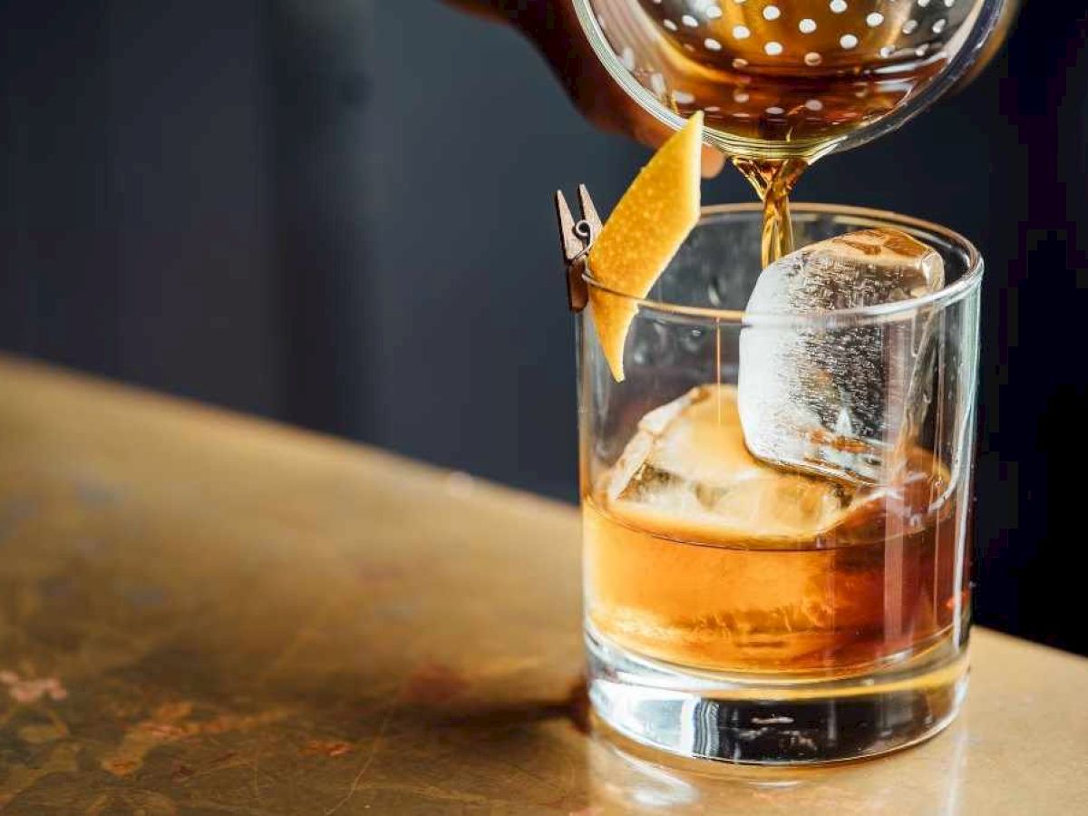 A drink is being poured into a glass with ice and a lemon twist, using a strainer for precision.