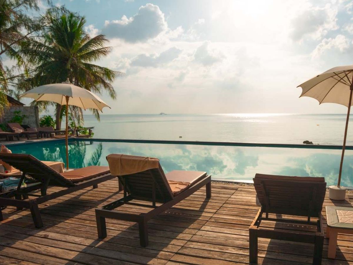 The image shows a serene seaside view with lounge chairs, umbrellas, and a pool overlooking the ocean, under a partly cloudy sky.