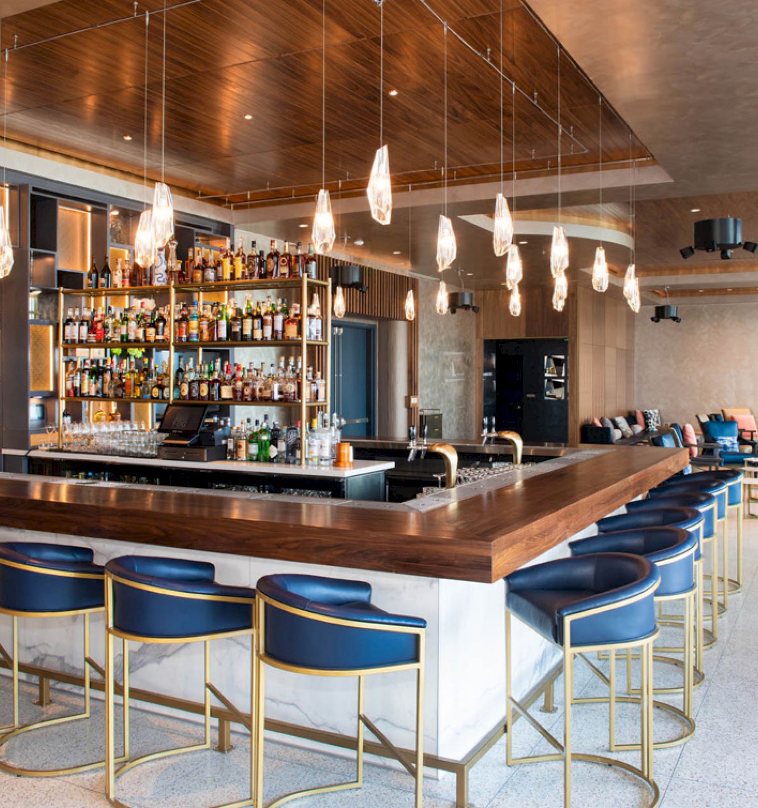 A modern bar area with stools, a well-stocked liquor shelf, hanging lights, and a cozy seating area in the background.
