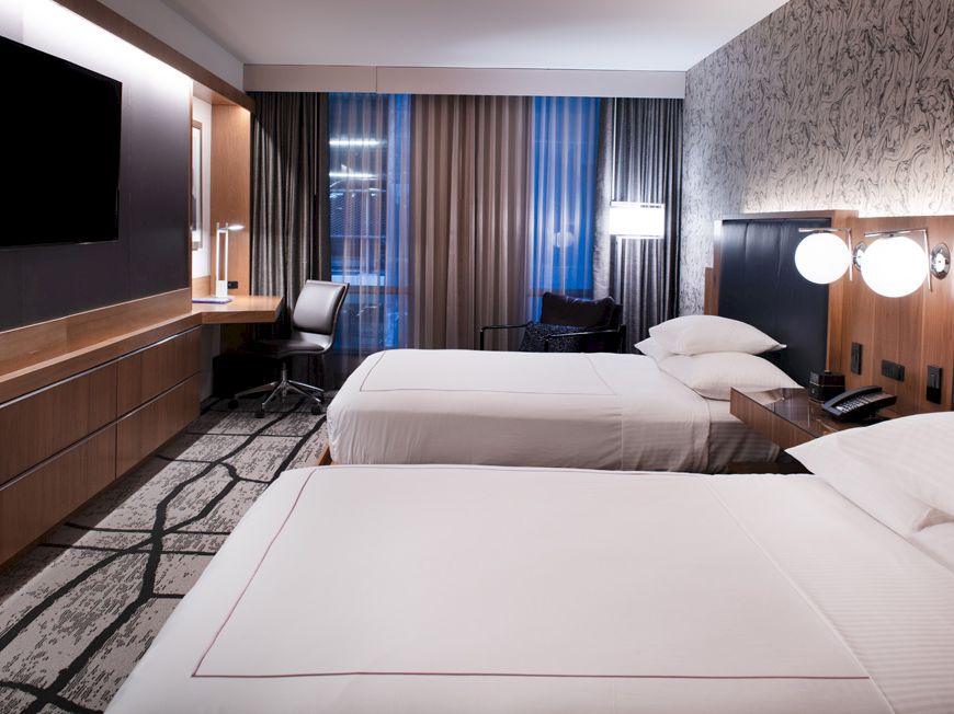 The image shows a hotel room with two beds, a desk with a chair, a wall-mounted TV, and stylish decor and lighting.