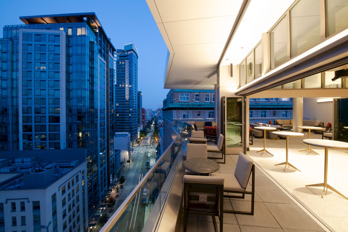 A modern balcony overlooking a city street at dusk with tall buildings and a sleek, furnished open-air area.