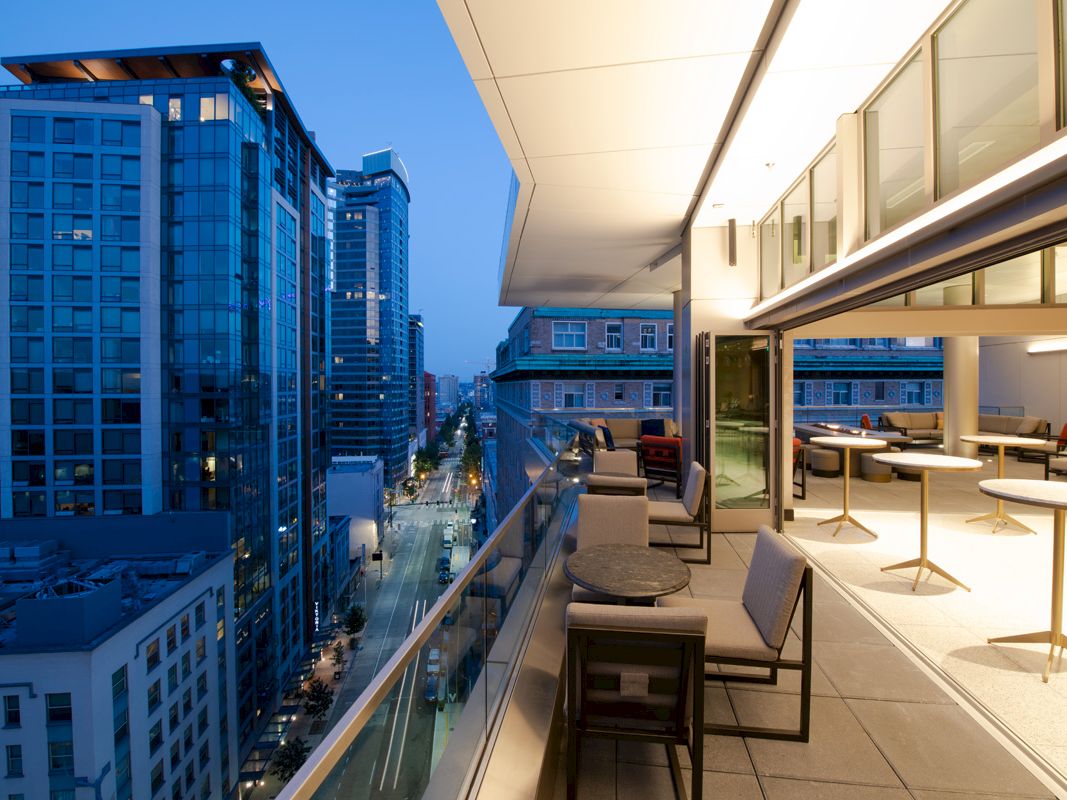 A modern balcony overlooking a city street at dusk with tall buildings and a sleek, furnished open-air area.