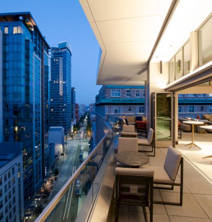 A modern cityscape at dusk with tall buildings, a lit street, and a sleek balcony restaurant featuring tables and chairs.