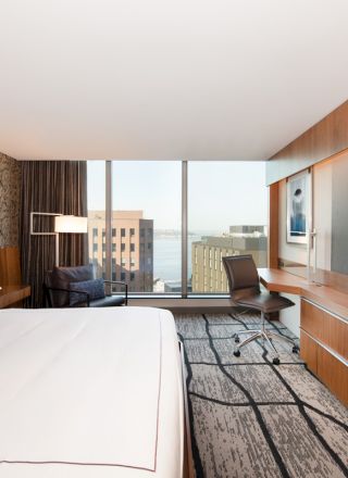 A modern hotel room with a large bed, a desk, a chair, a flat-screen TV, and a large window showing city and water views.