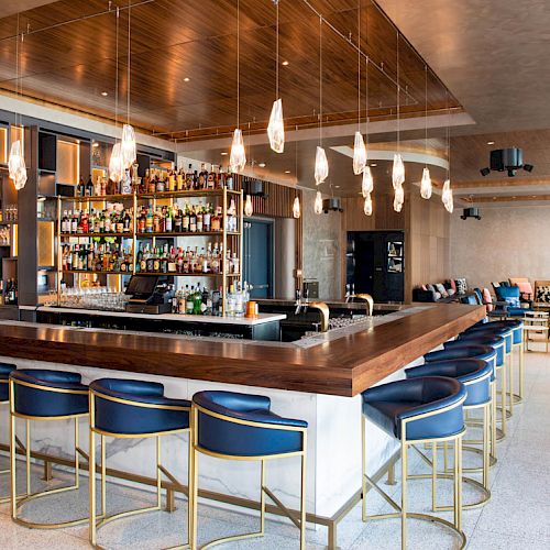 This image shows a modern bar with a stocked liquor shelf, stylish lighting, and blue barstools surrounding a wooden counter.