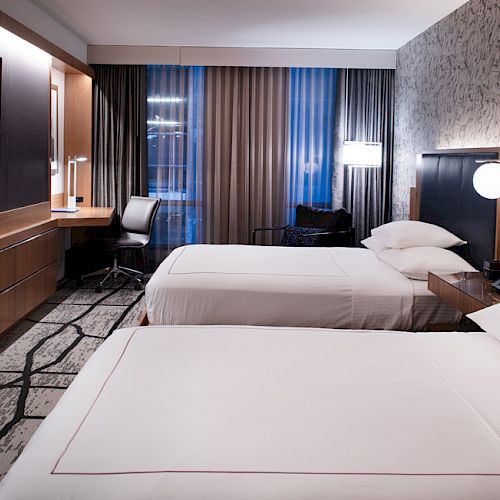 The image shows a modern hotel room with two beds, a flat-screen TV, desk, and ambient lighting, creating a cozy atmosphere.