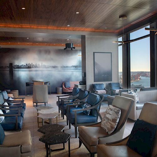 A modern lounge with armchairs, tables, and large windows overlooking a cityscape and water; skyline art decorates the wall.