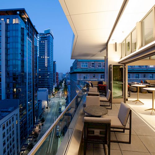 A modern balcony overlooking a cityscape at dusk, with tables and chairs, and illuminated buildings in the background.