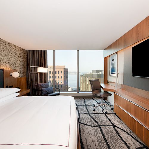 A modern hotel room with a large bed, desk, and TV, featuring a window with a city view and contemporary decor.